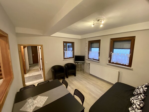 Ein gemütlicher Wohnraum mit Esstisch, zwei Stühlen, einer Couch, einem Fernseher und einem Fenster mit Vorhängen.