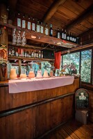 A rustic bar area with wooden shelves holding bottles and glasses, a table with pitchers of drinks, and a chalkboard menu.
