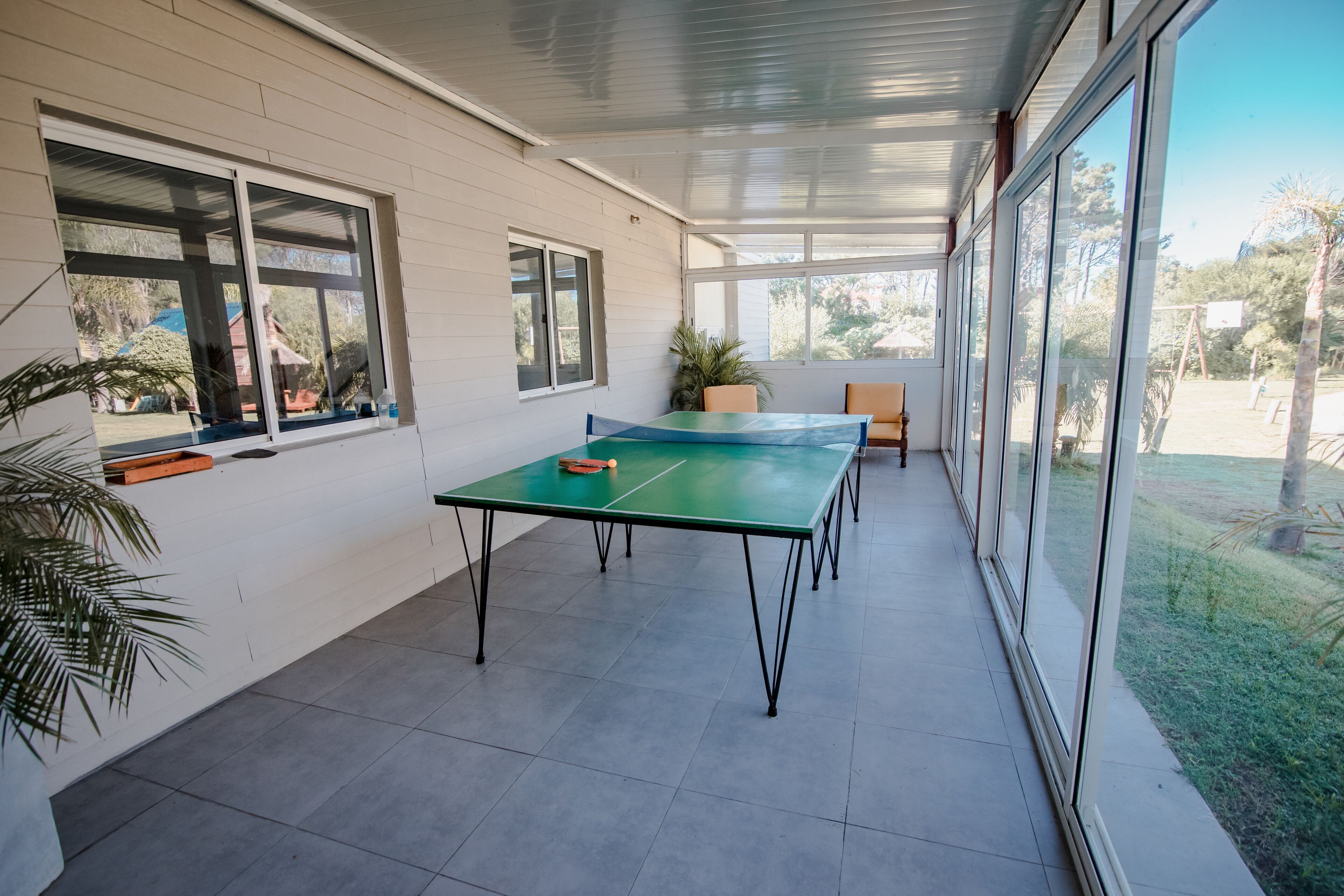Une terrasse couverte équipée d’une table de ping-pong, de plantes en pot et de grandes fenêtres offrant une vue sur l’extérieur.