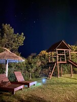 A night scene with a wooden play structure and lounge chairs under a thatched roof.