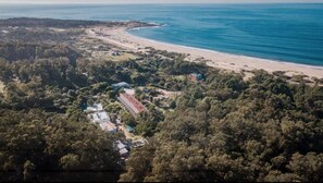 An aerial view of a coastal area with a beach, dense forest, and a few buildings.