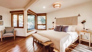 A bedroom with a large bed, wooden flooring, a bench, a nightstand, and a window with a view of trees. - Hidden Oasis Hot Tub-Disney Train Track. (Julian)