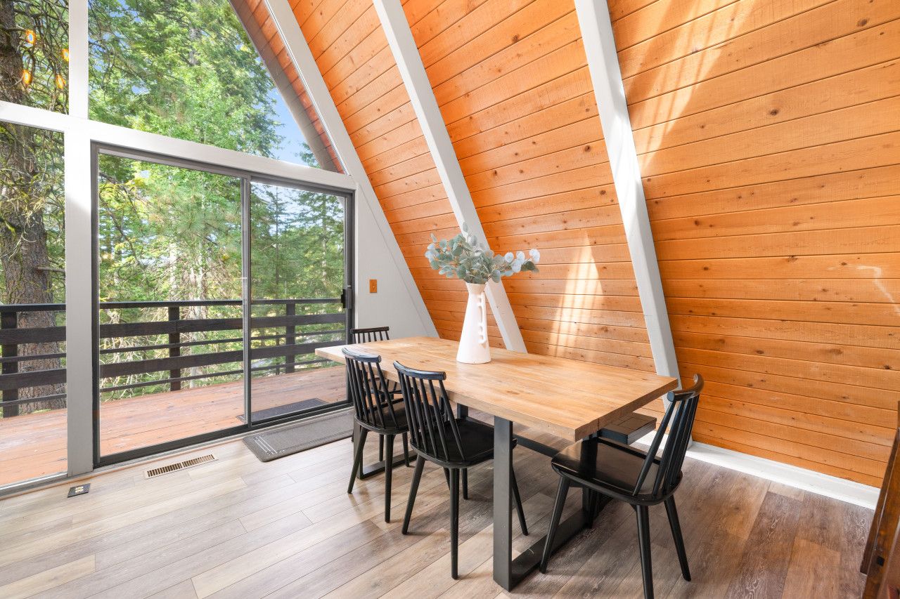 A dining area with a wooden table and chairs, large windows, and a view of the outdoors.