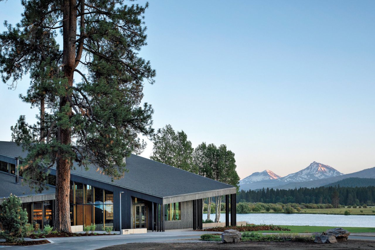 Un edificio moderno con ventanas amplias, un área exterior techada y vistas a un lago y montañas.