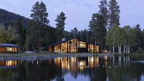 Un chalet moderne doté de grandes fenêtres, d’un étang aux eaux calmes et d’un paysage boisé en arrière-plan.