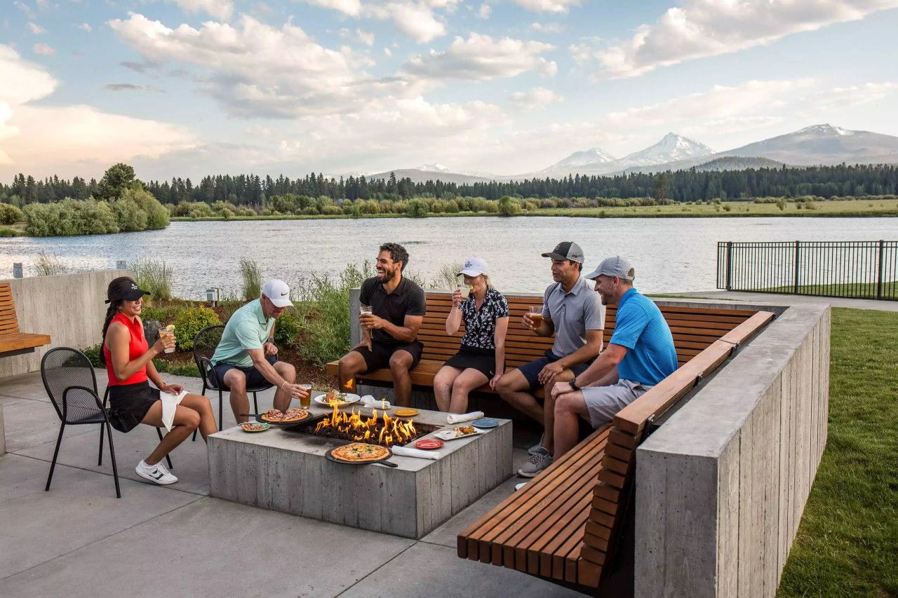 A group of people sitting around a fire pit with a lake and mountains in the background.