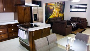 A modern kitchen with a central island, stainless steel appliances, and wooden cabinets. There is a dining area with a glass table and grey upholstered chairs. - Amplio departamento, con la mejor ubicación en la ciudad (Chihuahua)