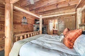 A wooden cabin bedroom with a bed, a fireplace, and a door leading to another room.
