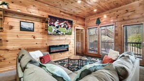 A cozy cabin interior with a wooden ceiling, a large TV mounted on the wall, a fireplace, a sofa with cushions, and a view of a staircase leading outside. - Stop Booking 2 Cabins. This One Sleeps 34. (Pigeon Forge)
