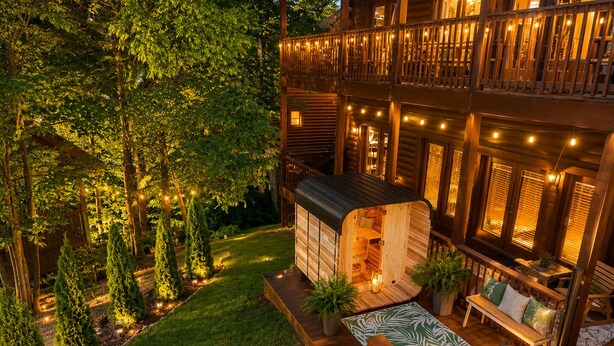 A wooden cabin with a deck, surrounded by trees and a well-lit garden. - Stop Booking 2 Cabins. This One Sleeps 34. (Pigeon Forge)