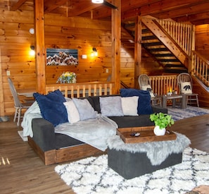 Interior de uma cabana de tronco com um sofá, cadeiras, uma escada e um quadro na parede.