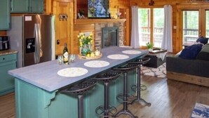 A wooden cabin interior with a kitchen, bar stools, a fireplace, and a sofa. - Stop Booking 2 Cabins. This One Sleeps 34. (Pigeon Forge)