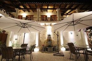 Un patio al aire libre con mesas y sillas bajo sombrillas, una fuente y piso de baldosas.