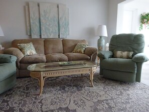 A living room with a brown sofa, a green armchair, and a glass coffee table. There are decorative pillows on the sofa and armchair.