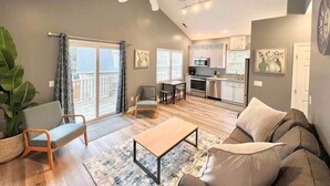 A spacious living area with a sofa, armchair, and coffee table, a balcony with curtains, a kitchen in the background, and a ceiling fan. - Charming, Beautiful, Treetop Loft, Prime Location (Wilmington)