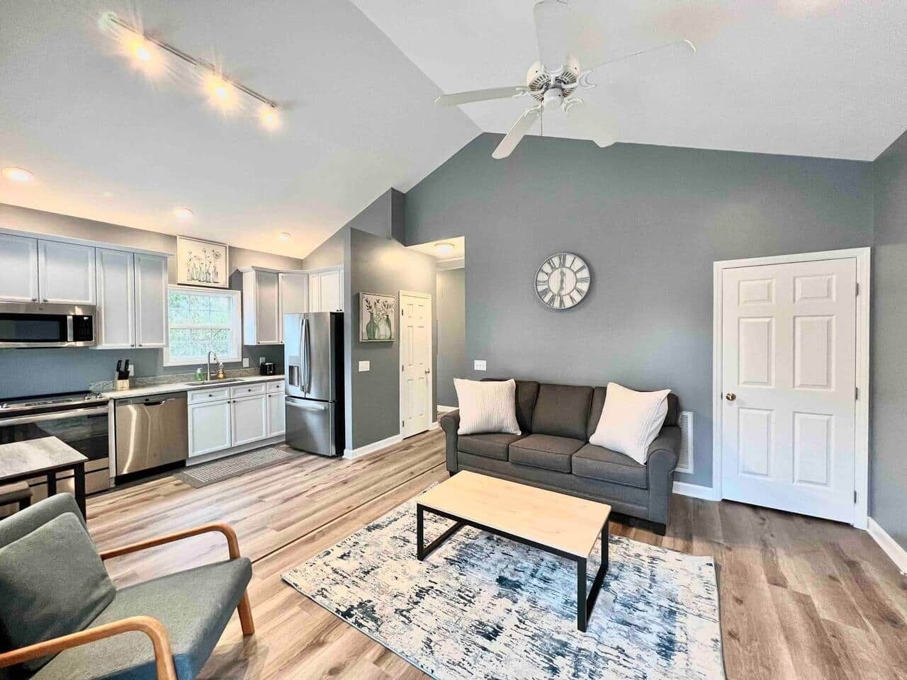 A modern kitchen with stainless steel appliances, a dining area with a table and chairs, a gray sofa, and a clock on the wall.