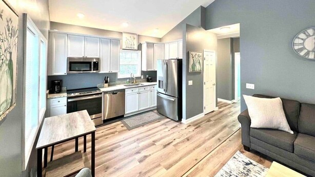 A modern kitchen with stainless steel appliances, a dining area with a table and chairs, and a living room with a sofa and a clock on the wall. - Charming, Beautiful, Treetop Loft, Prime Location (Wilmington)
