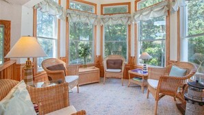 Une pièce chaleureuse, meublée de mobilier en osier, dotée de grandes fenêtres et offrant une vue sur la verdure.