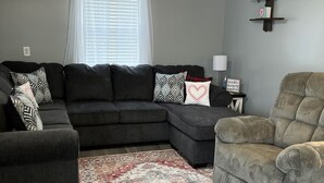 Ein Wohnzimmer mit Ecksofa, einem einzelnen Sessel, einem gemusterten Teppich, einem Deckenventilator und einem Fenster mit Jalousien.