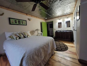 Un dormitorio con cama, mesita de noche, lámpara, una puerta verde, un baño con lavabo y espejo, y un ventilador de techo.