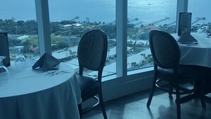 A dining area with a table set for two, large windows offering a view of a body of water and a distant landscape. - Beach&Pool Are Open! Stunning Newly Remodeled Oceanfront Condo on Daytona Beach! (Daytona Beach)