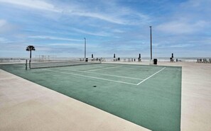 An outdoor tennis court with a net, surrounded by a fence, and a beach in the background.