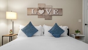 Een netjes opgemaakt bed met witte beddengoed en twee blauwe kussens, een bedlamp, een klein tafeltje met een bloevas, en een wanddecoratie waarop 'LOVE' staat met een hart in het midden.