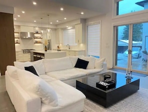 A modern living room with a white sectional sofa, a black coffee table, and a large window offering a view of the outside. - Luxury 5 BR home w/ pool perfect for a getaway. (Langley)
