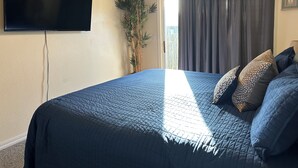 Un dormitorio con una cama, televisión montada en la pared, una planta y una ventana con cortinas.