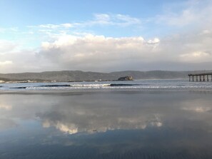 A beach with a pier extending into calm waters, a rocky outcrop in the distance, and a forested hill range.
