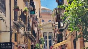A narrow street with buildings on either side, featuring balconies with plants and a prominent dome structure in the background. - Teatro Massimo Apartament self check-in (Palermo)