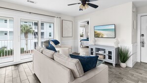 A spacious living room with a beige sofa, a ceiling fan, and a large window overlooking a balcony.