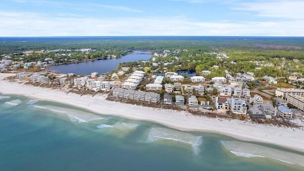 An aerial view of a coastal residential area with beachfront properties, a lake, and surrounding greenery.