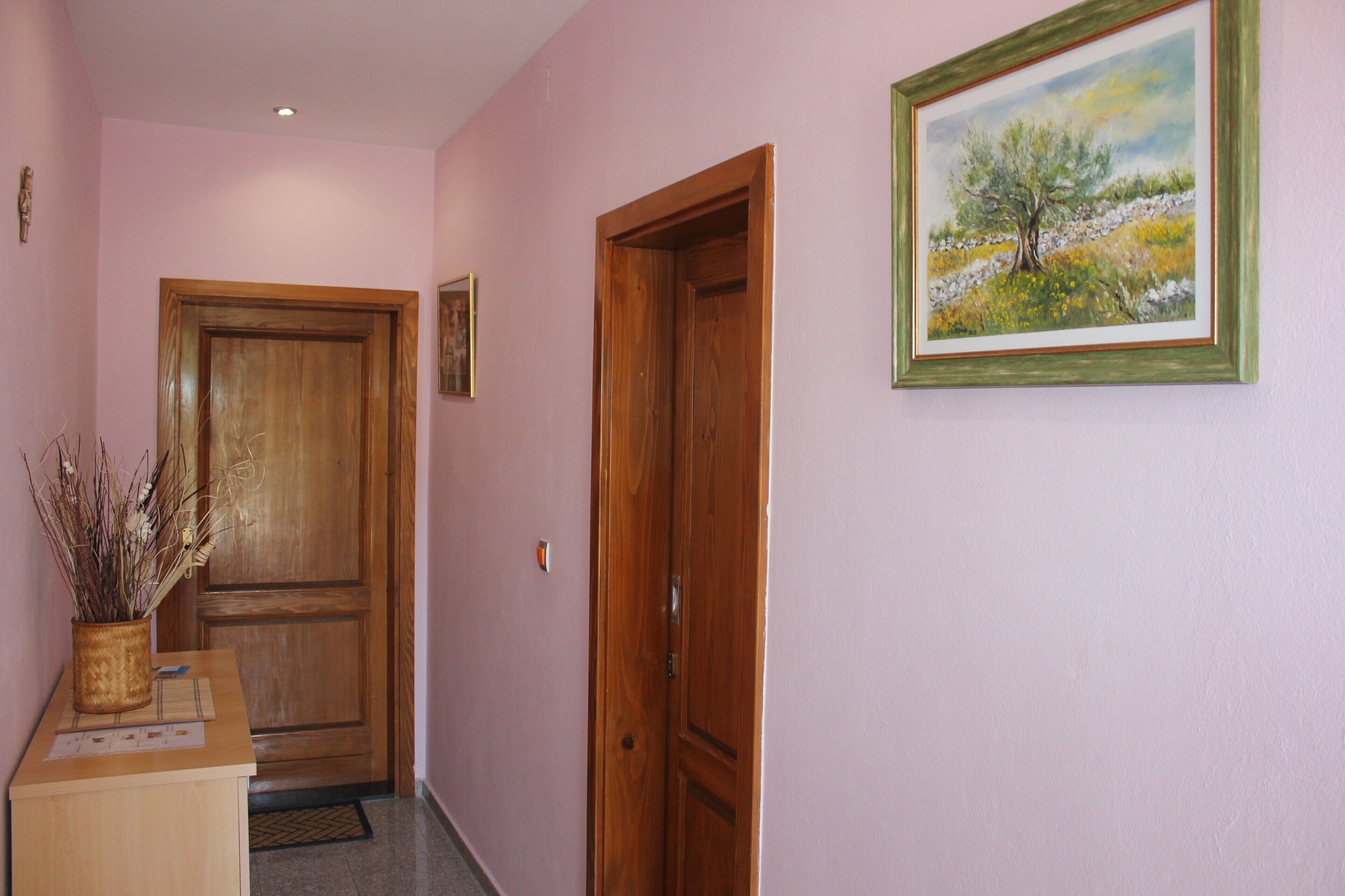 A hallway with a wooden door, a framed painting, and a desk with a vase of dried plants.