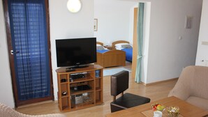 A hotel room with a television on a wooden stand, a dining table with a bowl of fruit, a sofa, and a bed with blue bedding.