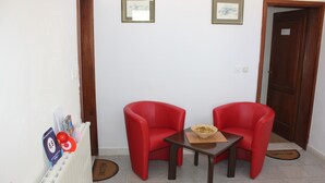 Ein Zimmer mit zwei roten Stühlen, einem kleinen Tisch und einer Heizung. An der Wand befinden sich gerahmte Bilder und eine Tür mit einem Schild.