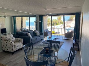 A spacious living area with a sofa, armchair, and dining table. Large windows provide a view of the outdoors.