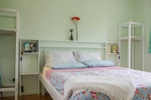 A bedroom with a bed, white shelving unit, and a small table with a vase and a red flower. - Charming Farm Cottage "Hibiscus" Breakfast & Bikes  (Matavera)