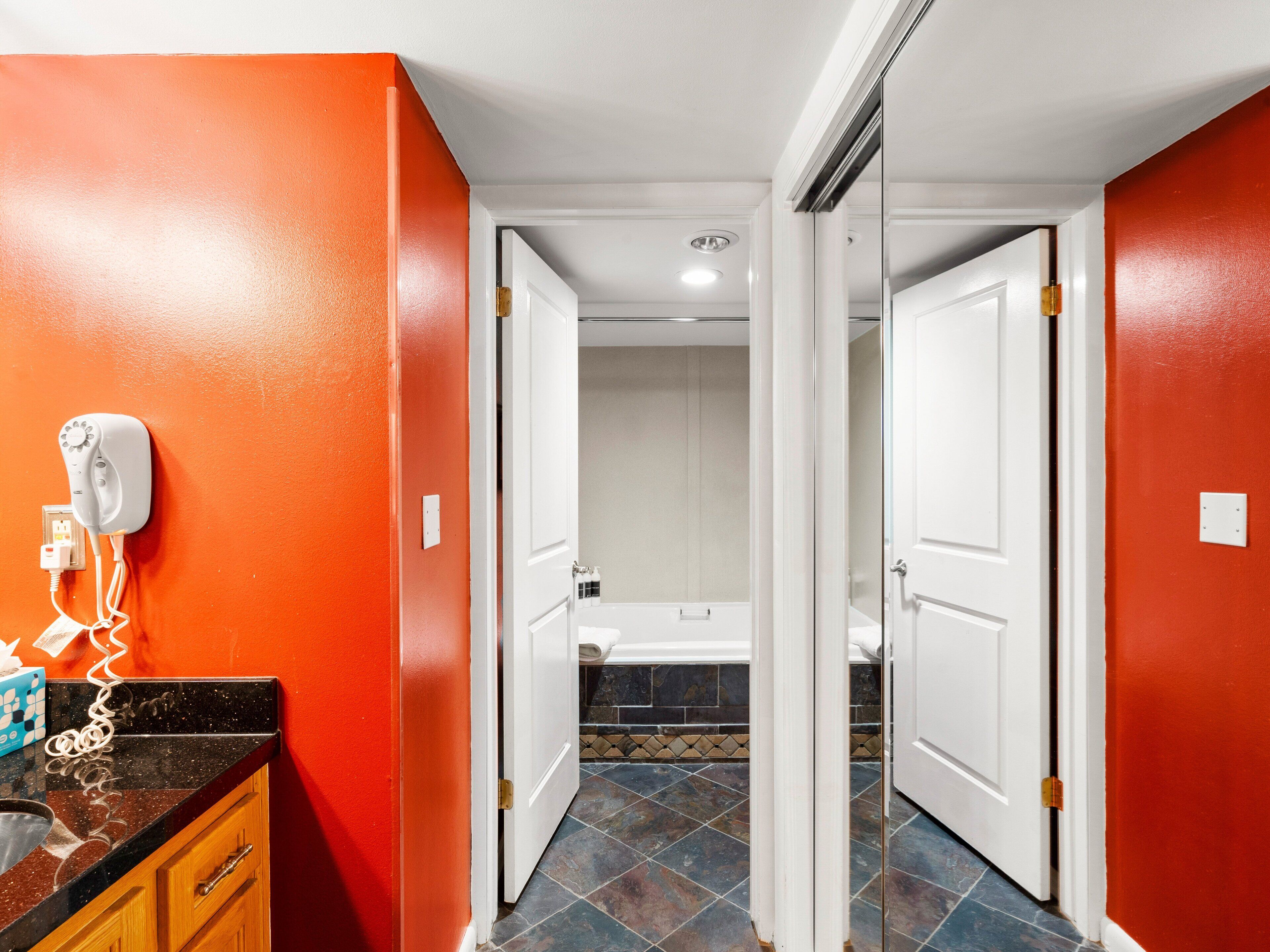 Un pasillo con una pared naranja, una puerta blanca que da a un baño con bañera, y un secador de pelo en la pared.