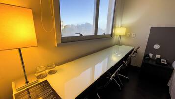 A hotel room with a long, white desk, two lamps, a mirror, and a view of the cityscape.