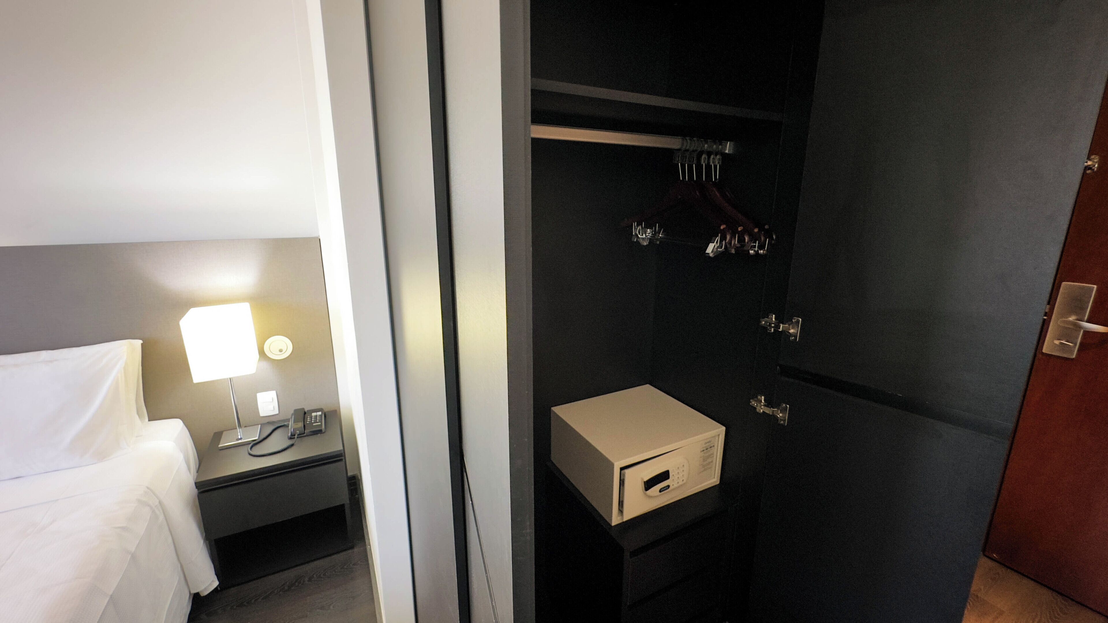 A hotel room with a bed, bedside table with a lamp, and a dark-colored wardrobe with a hanging rack.