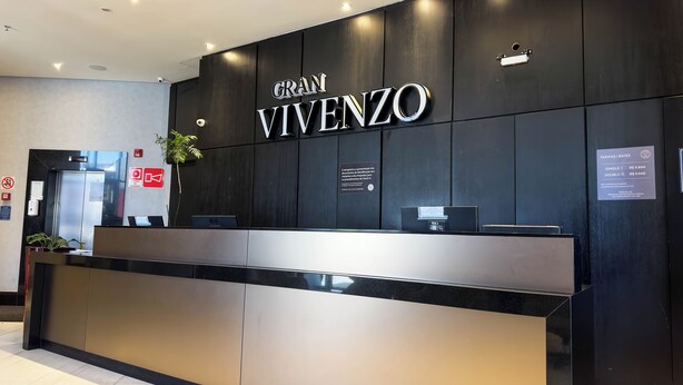 A hotel reception desk with a black and gold color scheme, a sign reading "GRAN VIVENZO", and a digital screen displaying rates. - Gran Vivenzo Cidade Nova (Belo Horizonte)