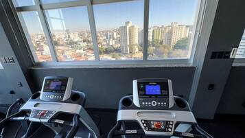 Duas bicicletas ergométricas com mostradores digitais em um quarto com uma janela grande com vista para a paisagem urbana.