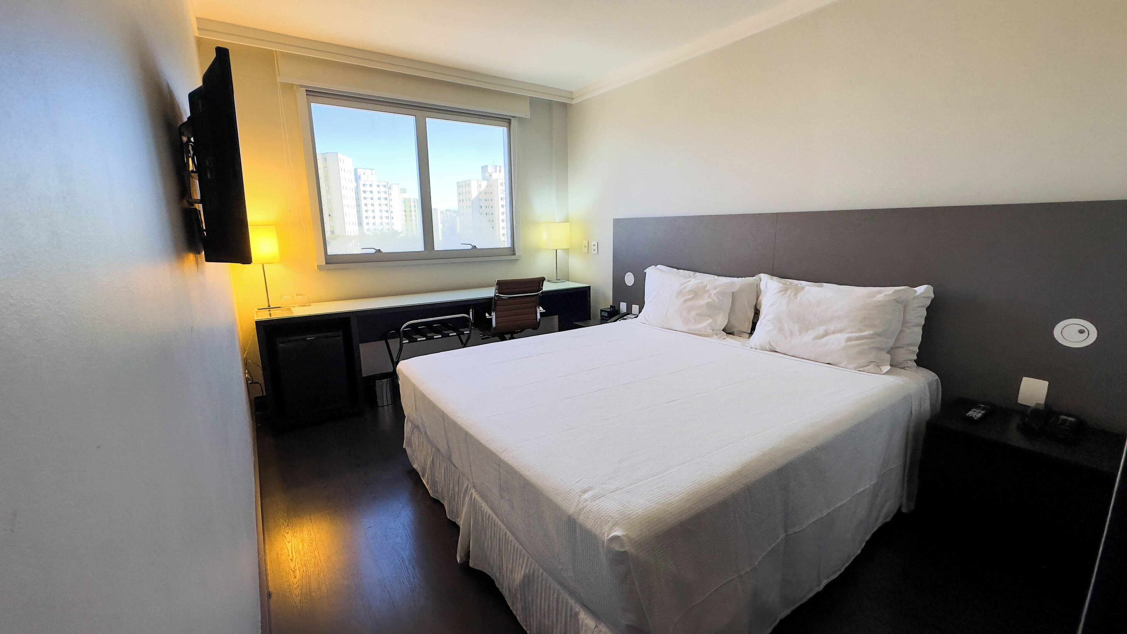 Habitación de hotel con una cama grande, un escritorio con silla, una ventana con vista a edificios y una mesita de noche con teléfono.