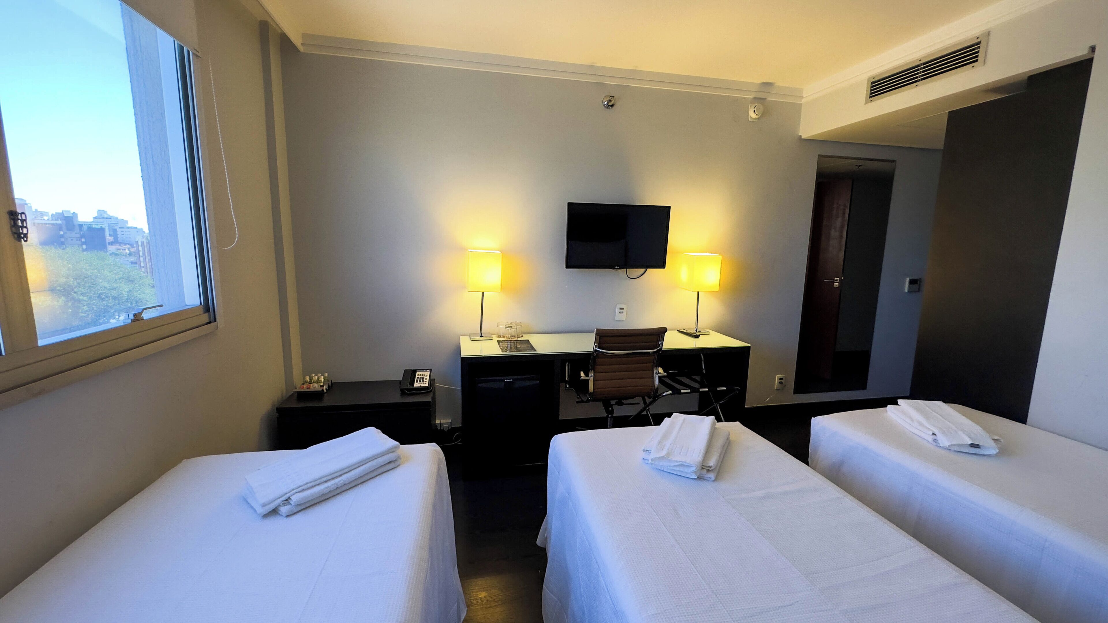 A hotel room with two beds, a desk, a chair, a TV, and two lamps.