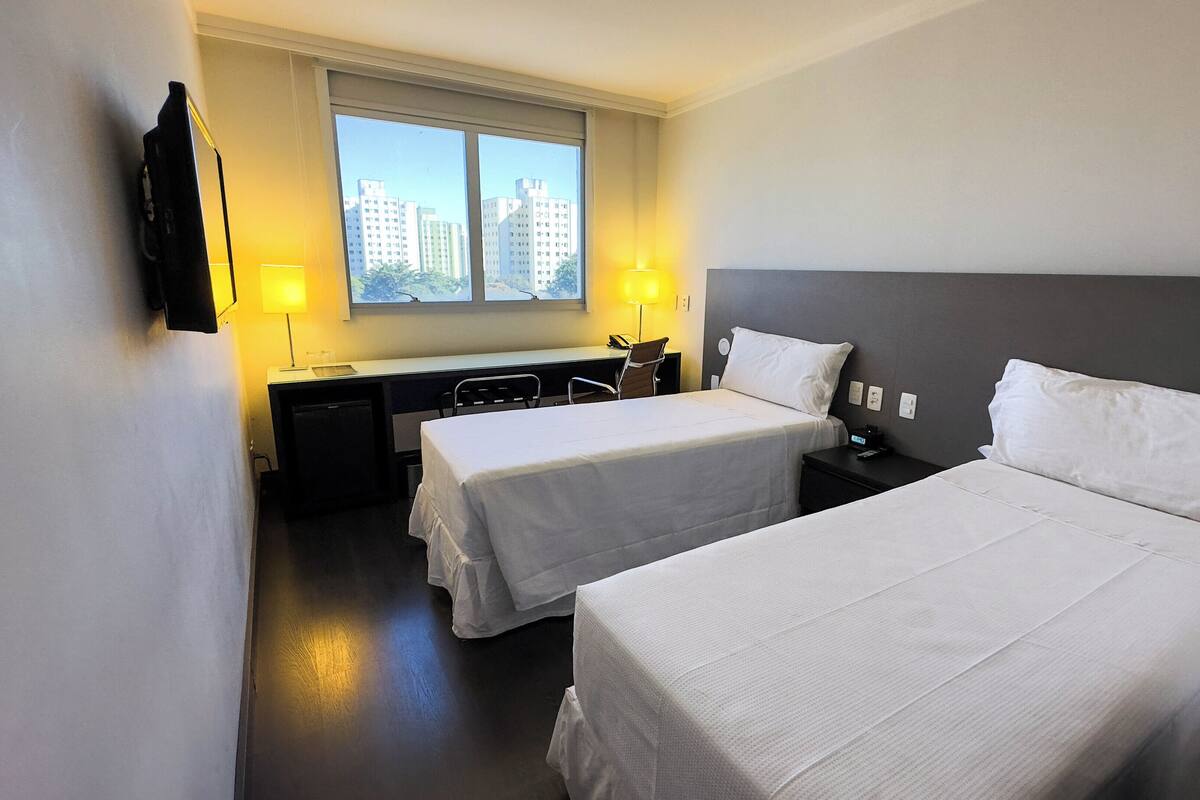 A hotel room with two single beds, a desk with a chair, a small TV, and a window with a city view.