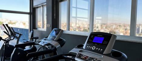 A gym with treadmills and a digital display, large windows, and a city view.