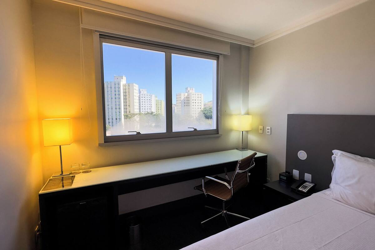 A hotel room with a bed, desk, chair, and a large window offering a city view.