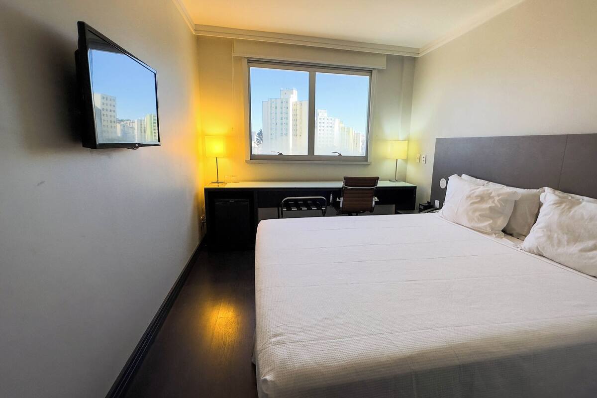 A hotel room with a large bed, a desk, a chair, a TV mounted on the wall, and a window with a city view.