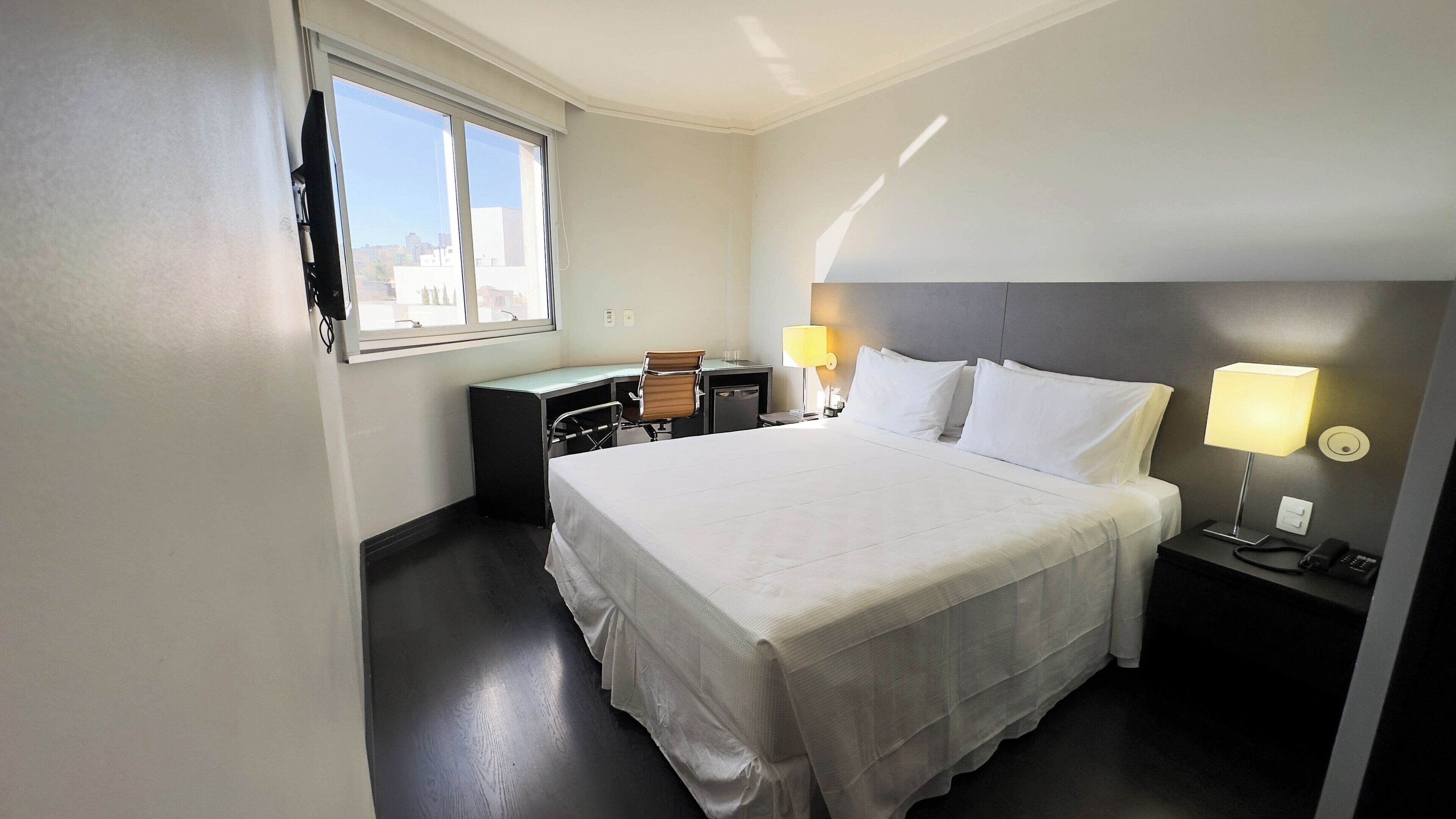 A hotel room with a large bed, a desk, a chair, two bedside lamps, and a window with a view of buildings.